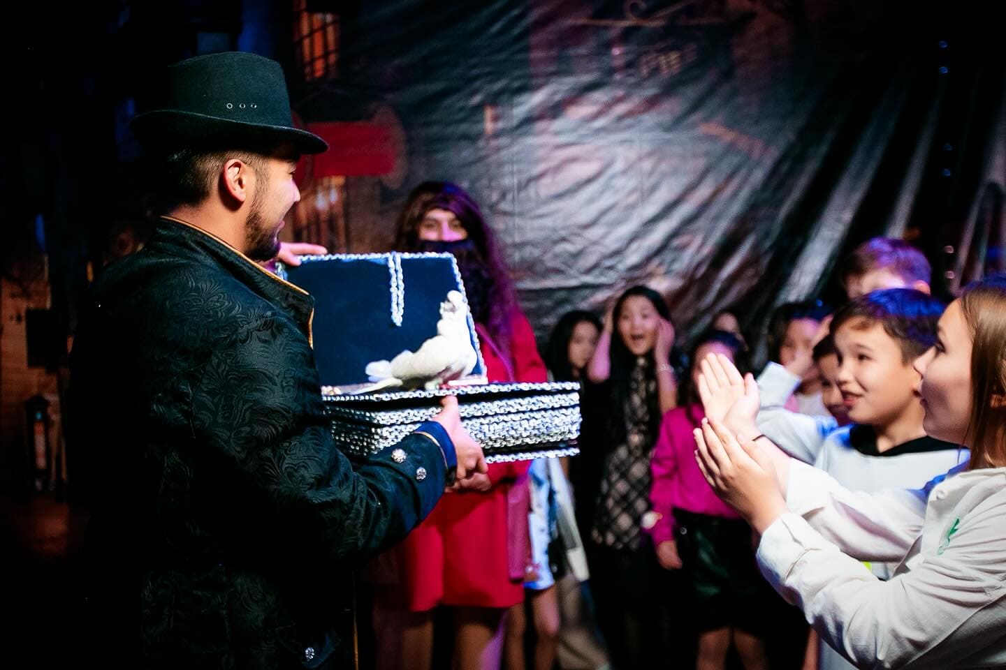 Фокусник в чёрной шляпе и темном костюме показывает трюк группе детей. Он держит открытую коробку, из которой выпрыгивает белый голубь, что вызывает восторг у детей. Некоторые дети хлопают в ладоши, а другие выражают удивление, прикрыв рот руками. Атмосфера полна магии и восхищения.