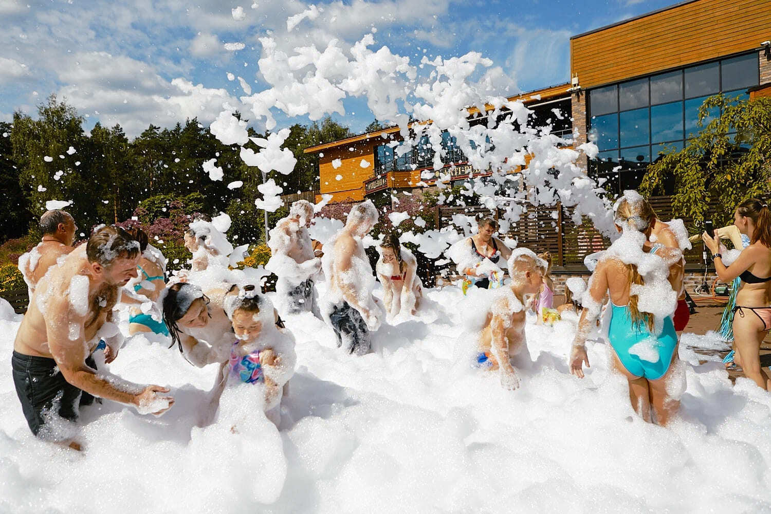 Группа людей, включая взрослых и детей, весело проводит время на пенной вечеринке под открытым небом. Все покрыты белой пеной, наслаждаясь игрой в мыльных пузырях на фоне солнечного дня и деревянного здания.