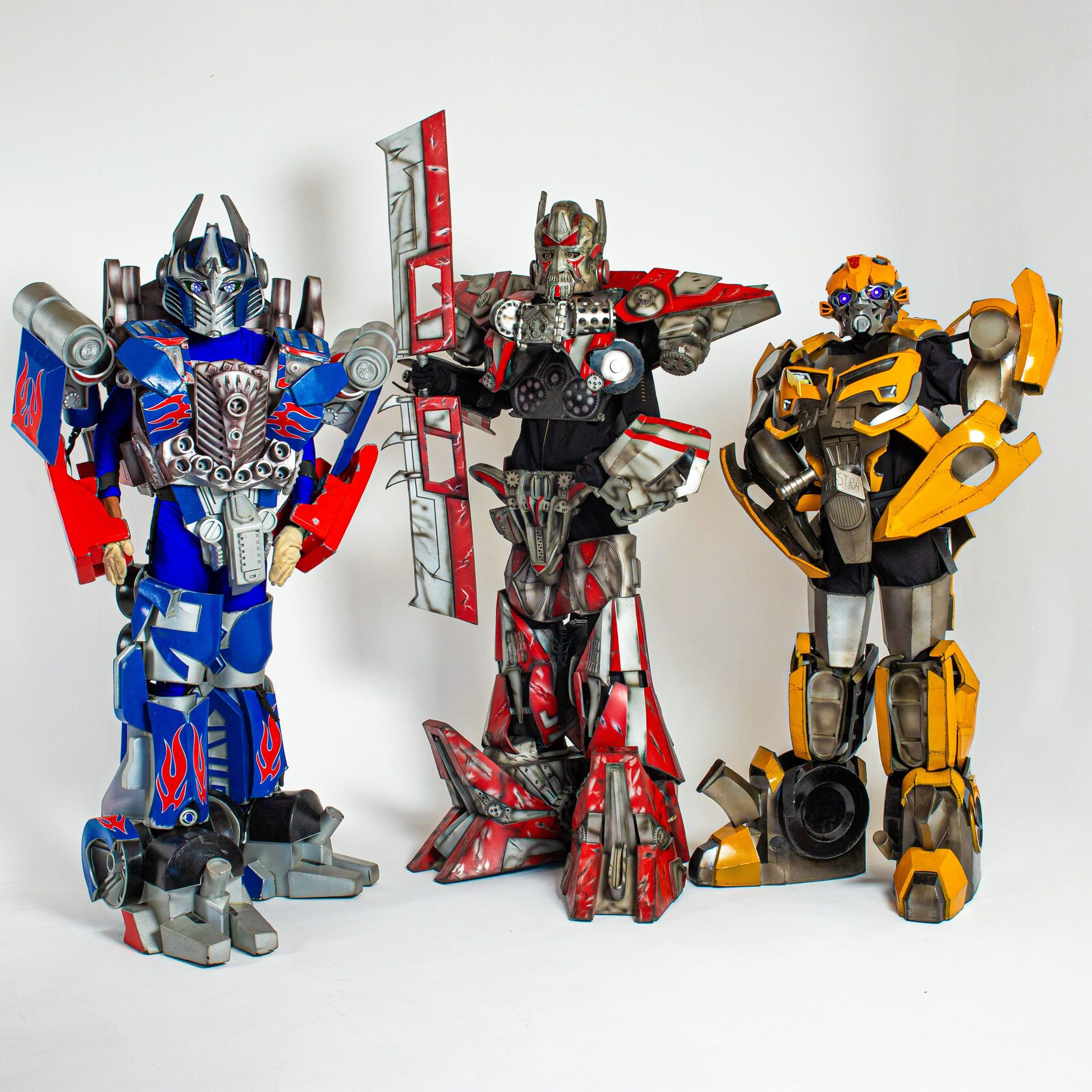 Три аниматора в костюмах трансформеров: слева Оптимус Прайм в синем и красном костюме с металлическими деталями, по центру персонаж в красно-серебристом костюме с мечом, справа — Бамблби в желтом и черном костюме.