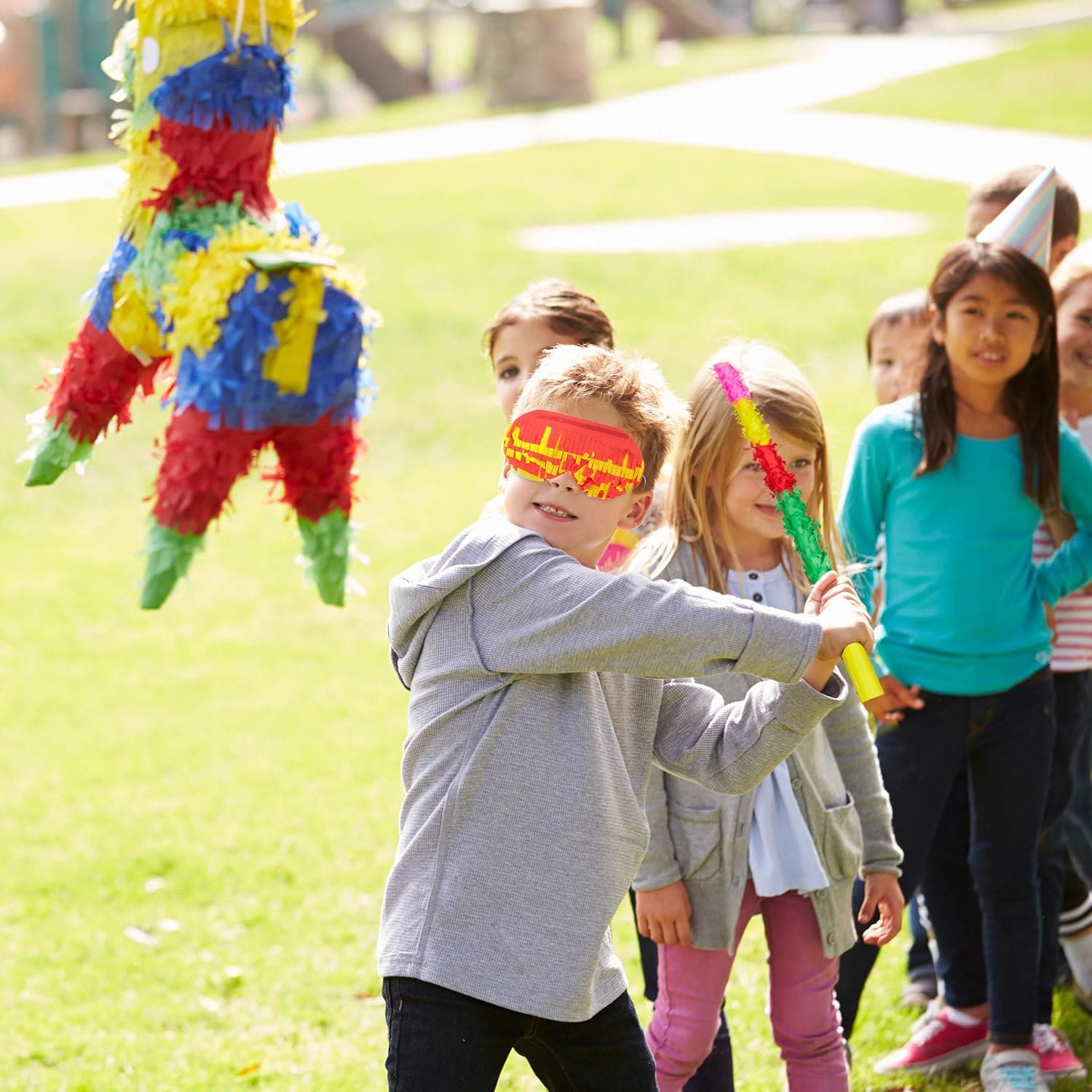 Мальчик с завязанными глазами бьет палкой пиньяту в виде разноцветного ослика, а за ним стоят дети, наблюдающие за игрой на зеленой лужайке.