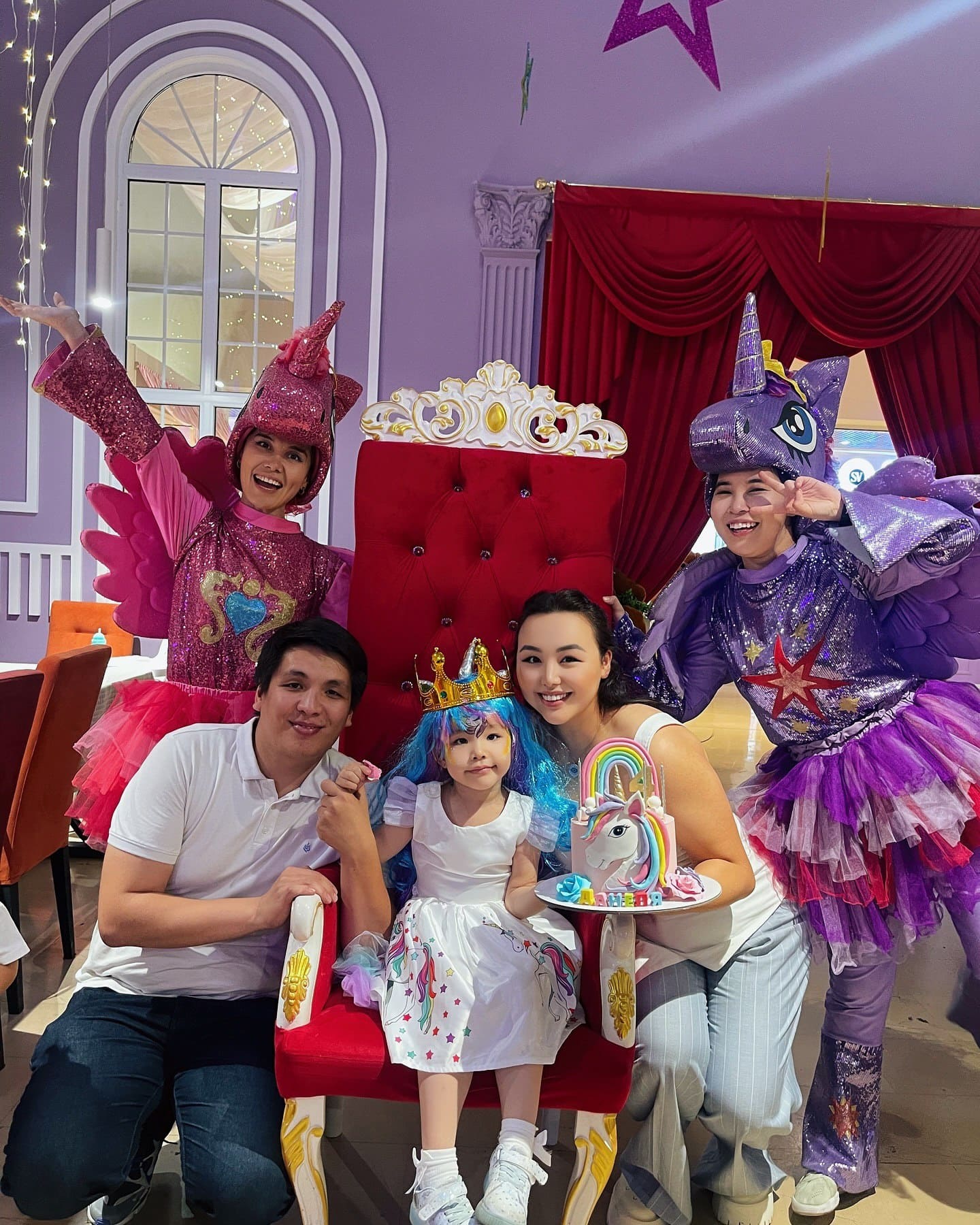 Маленькая девочка в белом платье с радужным принтом и синем парике сидит на красном троне, надев золотую корону. Рядом с ней сидят её родители, а по бокам стоят два аниматора в костюмах мифических существ — розового и фиолетового единорогов. Девочка держит праздничный торт с фигуркой единорога и радуги. Все улыбаются, создавая атмосферу радостного праздника.